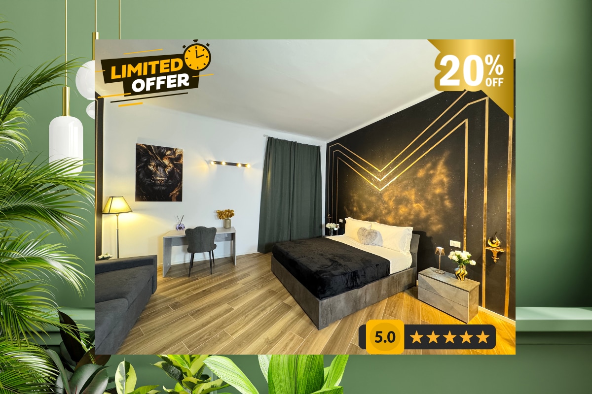 A modern bedroom features a spacious bed with dark bedding against a gold-accented wall. A small desk with a chair is situated to one side, complemented by a decorative arrangement of flowers. Soft drapes frame the window, adding a touch of elegance.