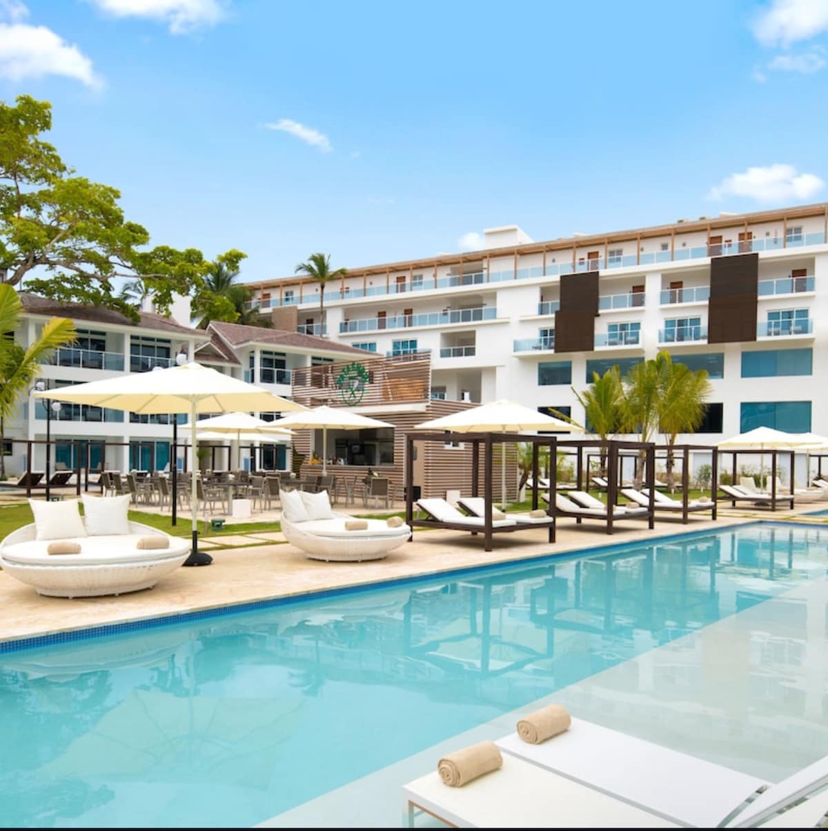 A spacious outdoor area is seen featuring a clear pool surrounded by lounge chairs and shaded umbrellas. A modern building stands in the background, complemented by palm trees and greenery, creating a relaxing environment for guests to enjoy.