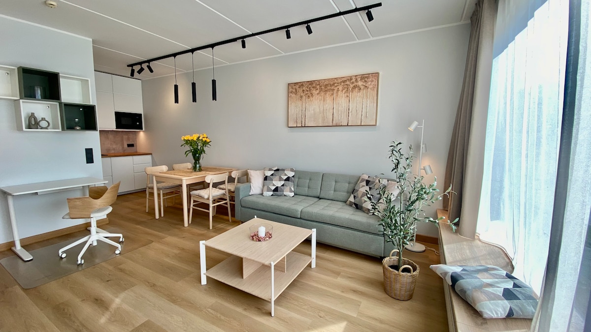 A bright and airy living space features a modern gray sofa adorned with decorative pillows. A wooden dining table is surrounded by matching chairs, while a small coffee table sits in front. Natural light floods the room through large windows, enhancing the minimalist décor.