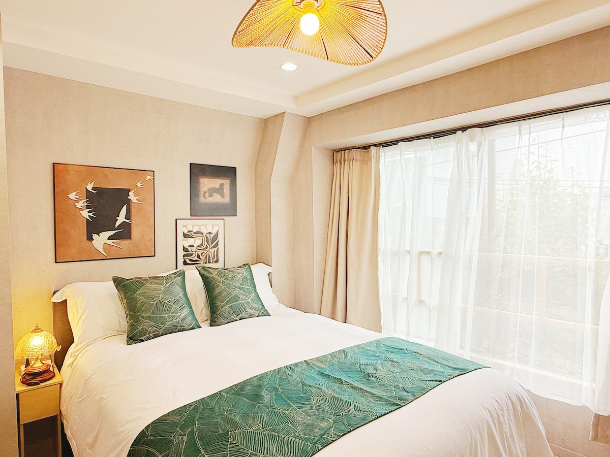 A cozy bedroom is presented, featuring a spacious bed adorned with a green patterned throw. Natural light enters through a large window, accentuated by sheer curtains. Art pieces are placed on the wall, complementing the warm, neutral tones of the room.