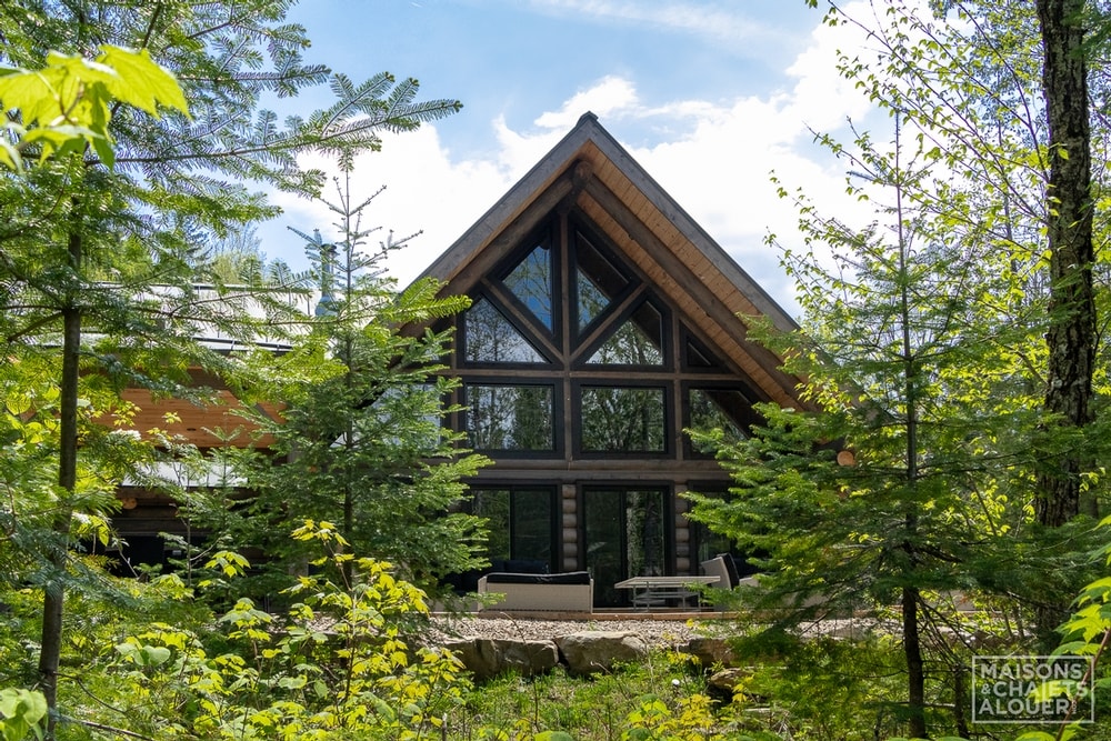 A modern log cabin is framed by tall trees, featuring a striking triangular roof and large windows that reflect natural light. An inviting outdoor seating area is visible, situated on a gravel patio. The surroundings include lush greenery, enhancing the secluded atmosphere.