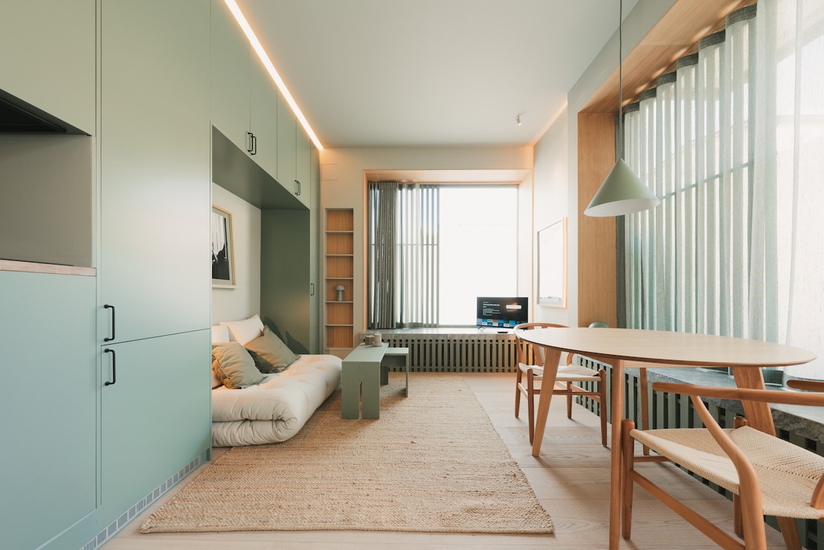A thoughtfully designed living area features soft seating with neutral tones, complemented by a small green coffee table. A wooden dining table with light chairs is positioned nearby. Natural light filters through sheer curtains, enhancing the calming atmosphere in the spacious interior.