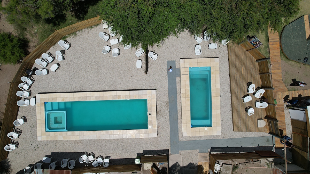 An aerial view showcases two swimming pools surrounded by a minimalistic outdoor area. Lounge chairs are arranged around the pools, which feature varying depths and textures. The landscaped environment includes gravel and wooden decking, creating a serene atmosphere ideal for relaxation.