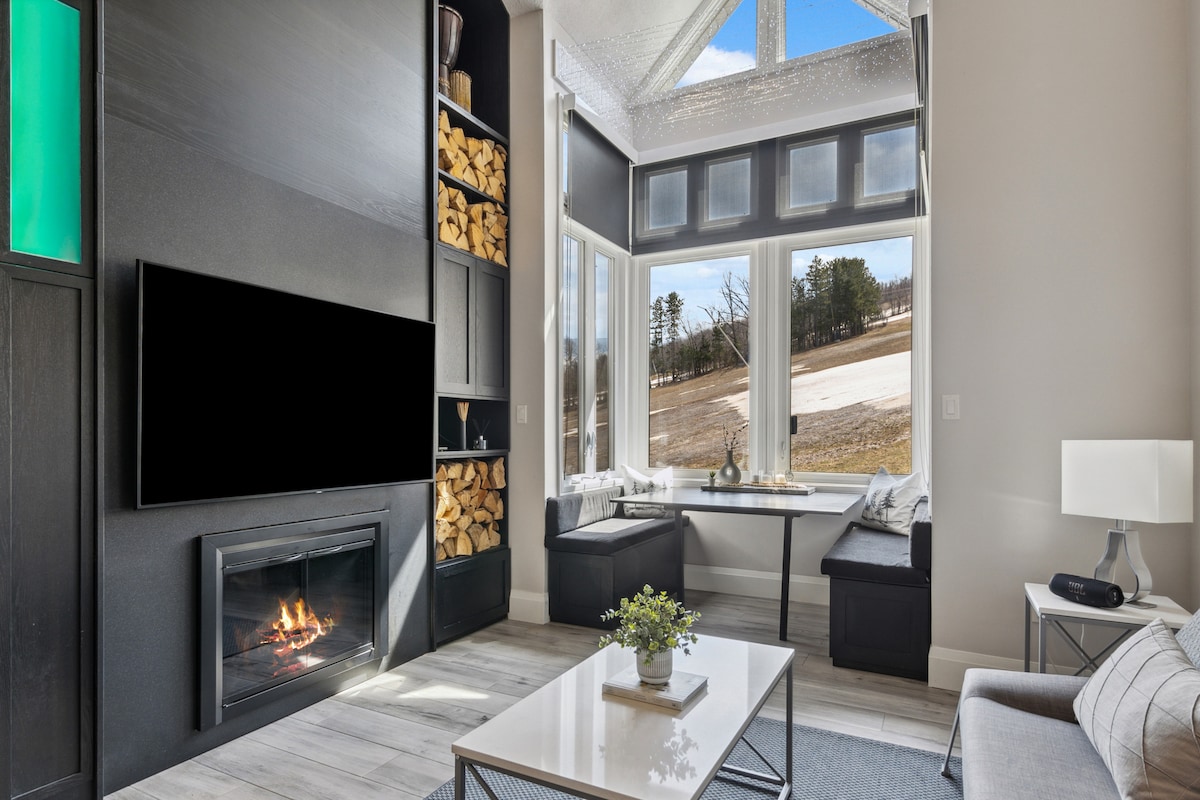 A cozy living area features cathedral ceilings and large windows framing views of the outdoors. A wood-burning fireplace is situated beneath a flat-screen TV, while a stylish coffee table rests on the floor. Built-in shelving holds neatly stacked firewood and decorative elements.