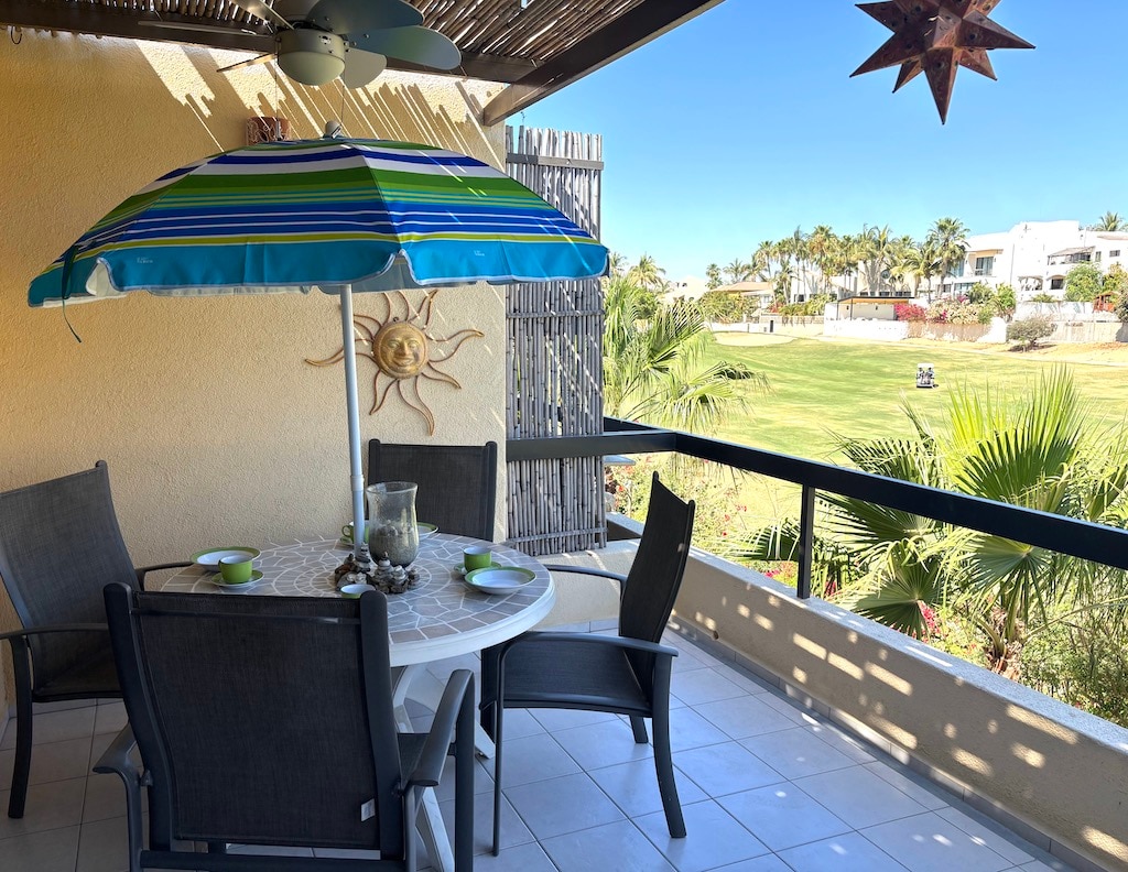 A balcony is featured with a round table surrounded by four chairs, topped with a colorful umbrella. A decorative sun motif is visible on the wall, while a view of green fairways and palm trees extends beyond the railing, against a clear blue sky.