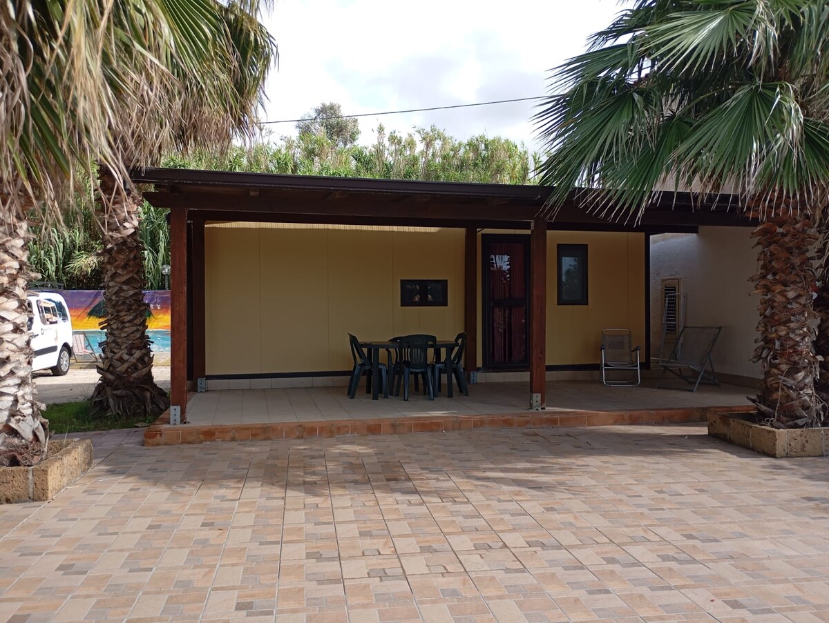 A shaded outdoor seating area is presented, featuring a table with four chairs under a wooden awning. Flanking the space, palm trees add greenery. The bungalow's exterior walls are painted in neutral tones, and a colorful mural is visible in the background.