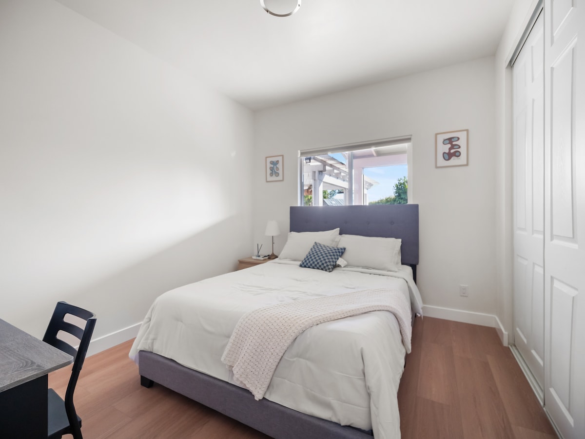 A private room features a spacious bed with soft linens and a decorative throw pillow. Natural light enters through a large window, illuminating the light walls. A desk and chair are positioned nearby, providing a work area. A closet is visible along one side.