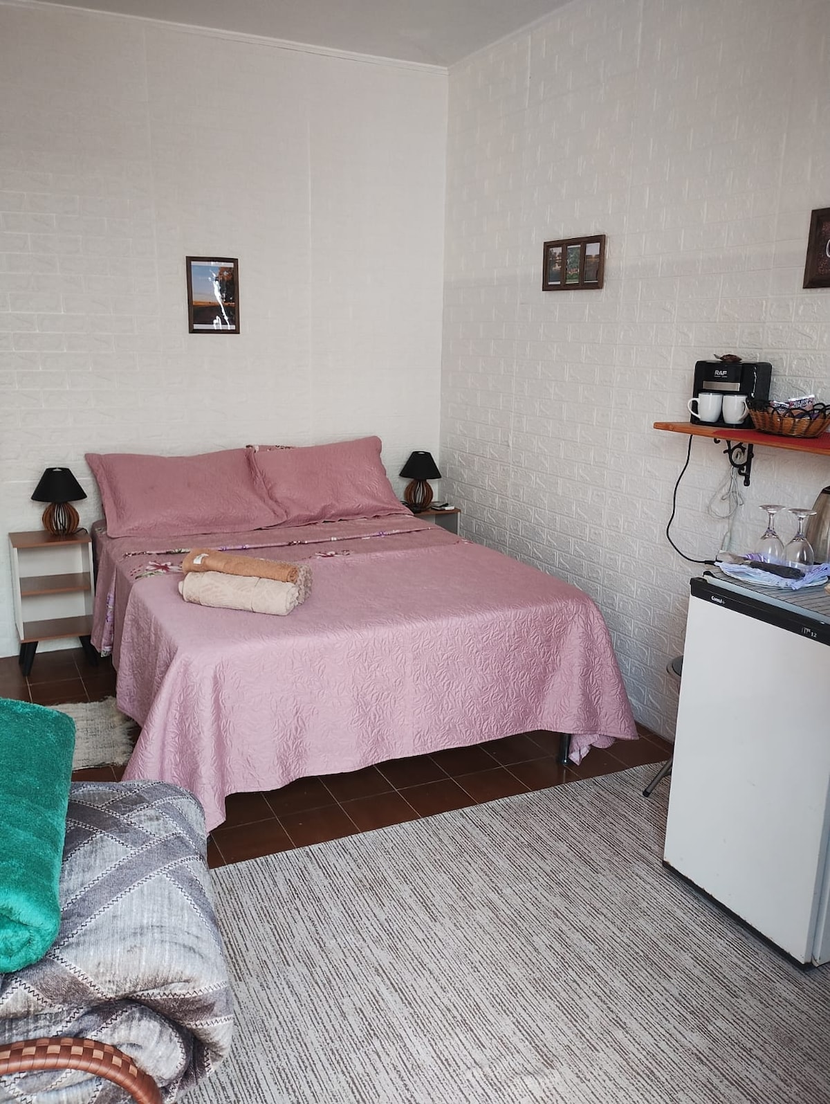 A compact sleeping area is arranged with a spacious bed covered in a light pink blanket. Nightstands are positioned beside the bed, each topped with a small lamp. A mini-fridge and a coffee maker are on a shelf against the wall, providing convenience for guests.