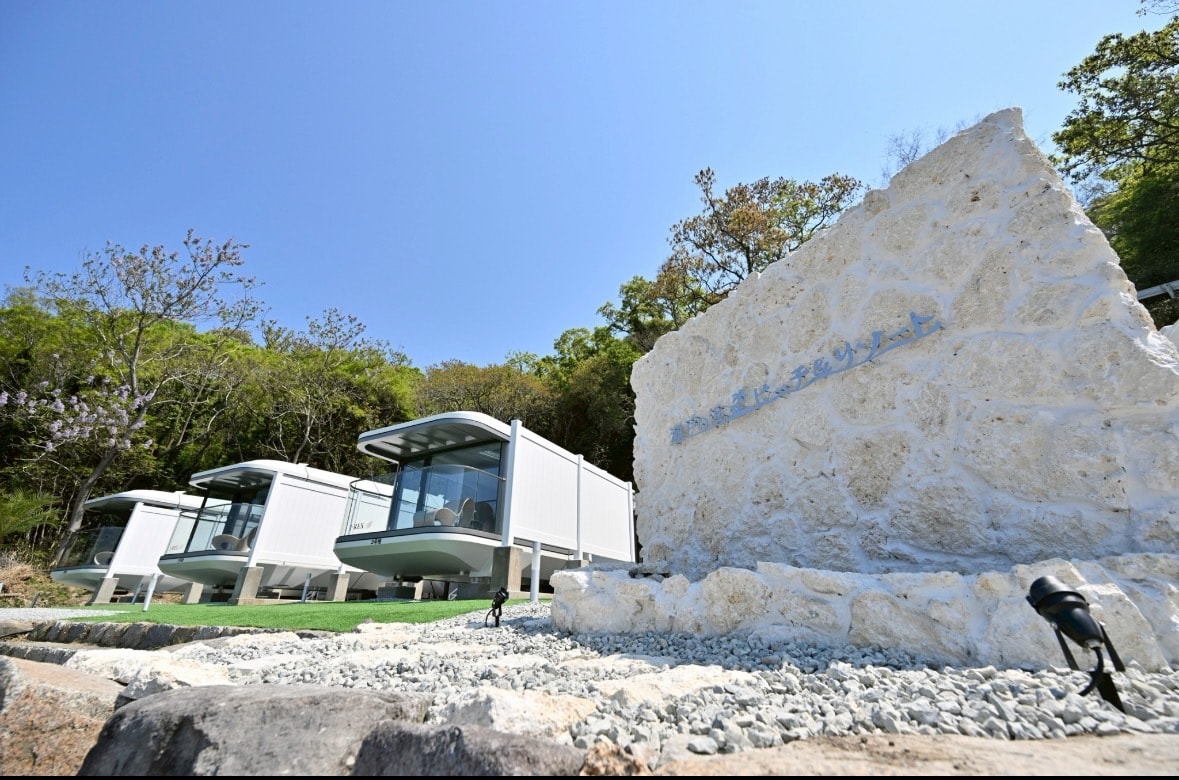 A modern container hotel is featured, showcasing multiple units set amidst lush greenery. A decorative stone wall with a blue inscription is positioned prominently in the foreground, complemented by a gravel path. Natural surroundings enhance the serene environment.