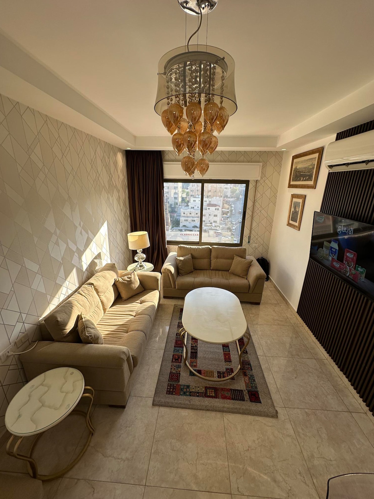 A well-lit living area features a neutral-toned sofa set and a round marble coffee table, complemented by a decorative area rug. Natural light enters through a large window, while a stylish chandelier adds contemporary charm above. An entertainment center with a flat-screen TV is positioned opposite the seating.