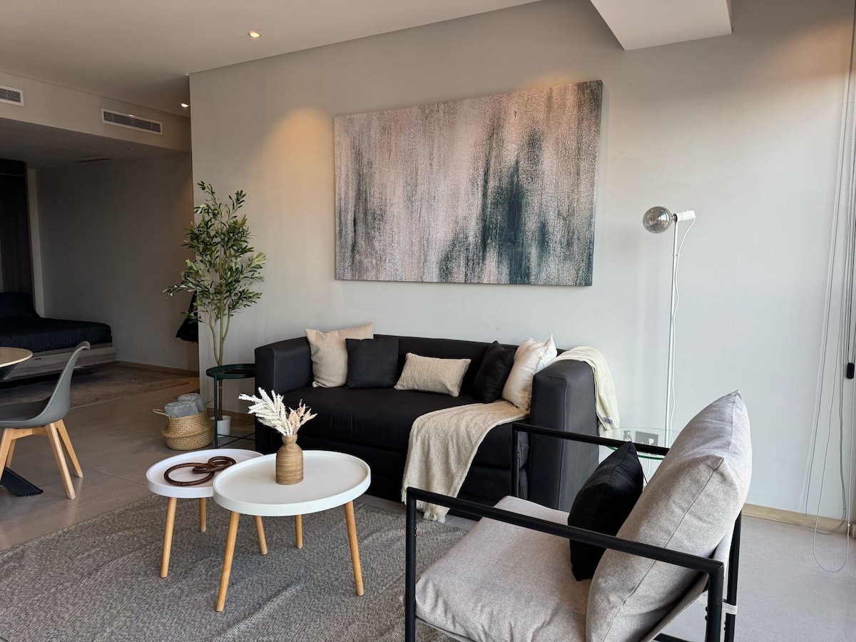 A modern living area features a dark sectional sofa adorned with light pillows, complemented by a soft throw. A sleek coffee table sits in front, surrounded by two chairs. A large artwork hangs above, while natural light enters through expansive windows, enhancing the spacious feel.