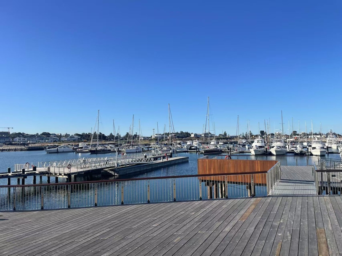 A serene marina view showcases numerous boats docked at wooden piers, reflecting in calm waters under a clear blue sky. The wooden boardwalk extends into the scene, providing access to the marina and enhancing the tranquil atmosphere of the surroundings.