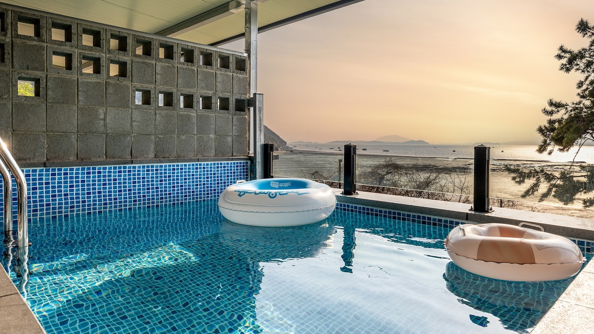 A private pool is surrounded by tiled decking, offering views of the ocean at sunset. Two inflatable tubes float on the calm water, while a stone wall provides privacy. The gentle hues of the sky reflect on the water's surface, creating a serene atmosphere.