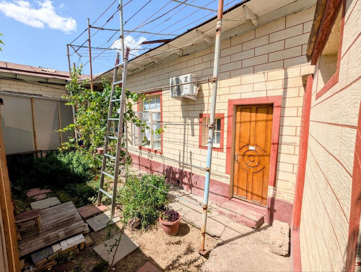 A small private yard is depicted, featuring a layout of stone tiles and greenery. An entrance door with a circular design is visible, and an air conditioning unit is mounted on the exterior wall. A ladder is set against the building, suggesting a maintenance area.