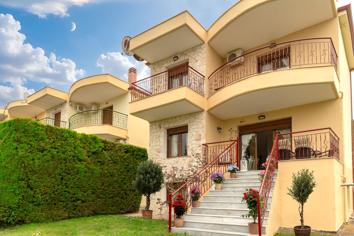 The villa features a charming exterior with a stone facade and decorative balconies. A set of stone steps leads to a welcoming entrance, surrounded by neatly trimmed greenery and planters filled with colorful flowers. A soft sky and crescent moon offer a serene backdrop.