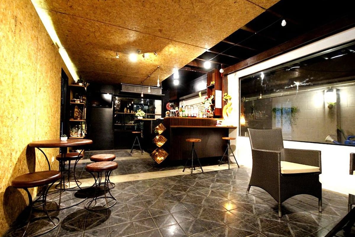 An inviting bar area features a long counter with a selection of bottles displayed on shelves. Several round tables with chairs are arranged around the space, complemented by sleek lighting and a stylish floor design. Large windows allow natural light to fill the area.