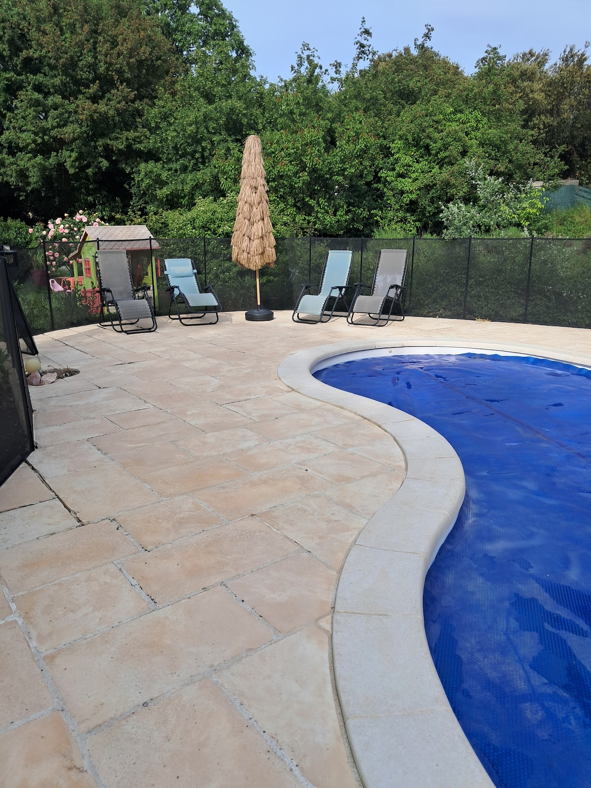 A shared swimming pool is framed by a paved terrace, featuring four lounge chairs arranged for relaxation. A tall parasol provides shade nearby, with greenery visible in the background, contributing to a serene outdoor setting.