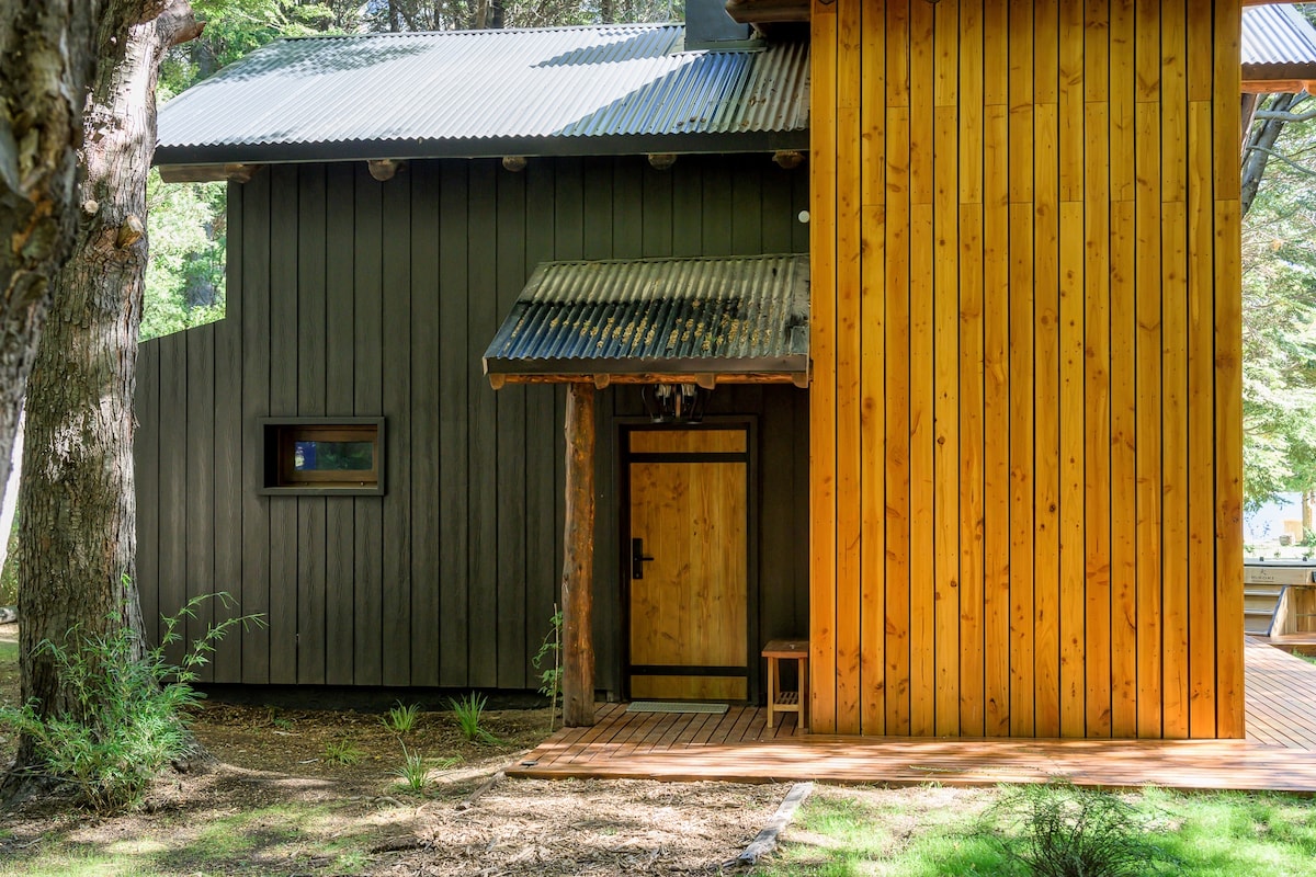The exterior of the cabaña is showcased, featuring a contemporary design with contrasting wood tones. The entrance is framed by a wooden door, surrounded by natural landscaping and wooden decking that leads to the interior. A rustic charm is present in the corrugated metal roof.