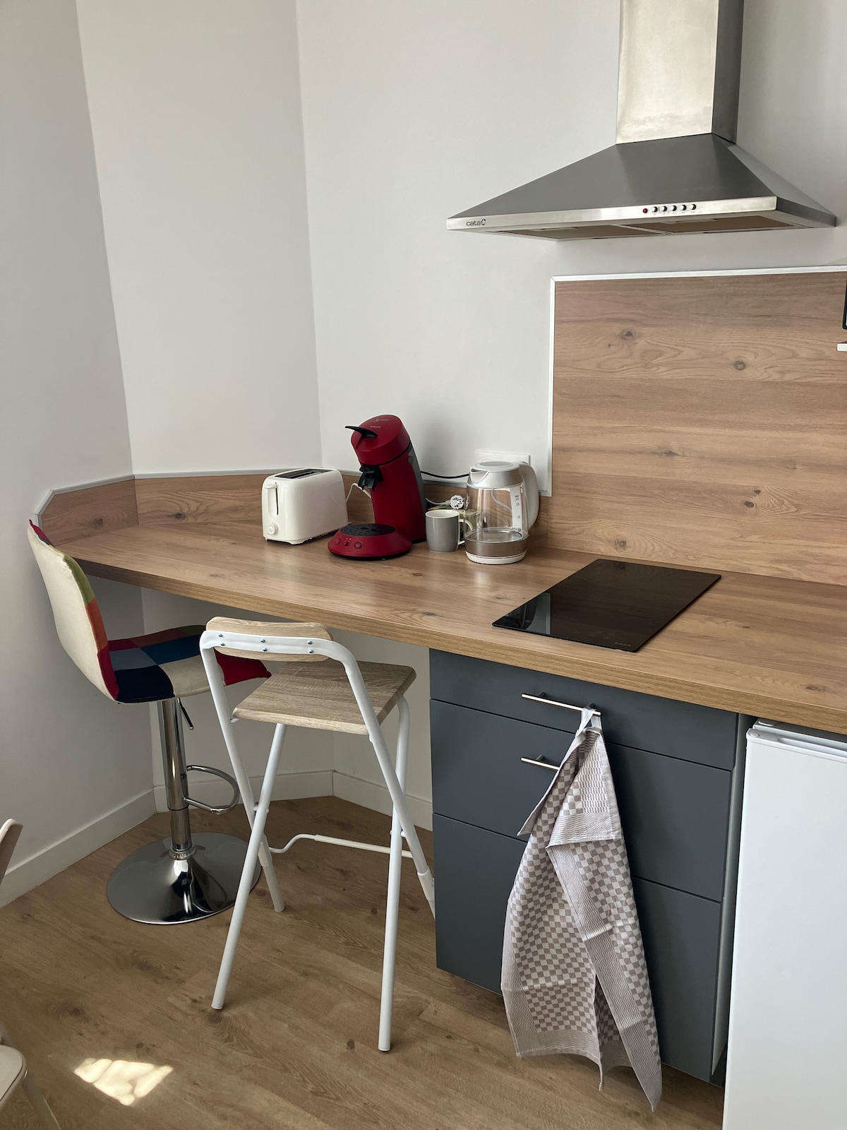 A compact kitchen area features a wooden countertop with a modern design. A bar stool with a colorful seat is positioned nearby. Kitchen appliances include a coffee maker, kettle, and toaster, all situated above a cooking surface. Natural light highlights the space.