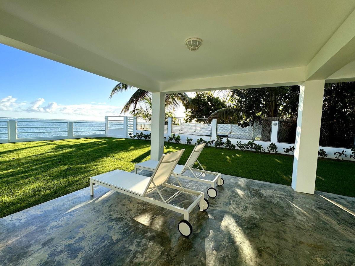 A serene outdoor space features three white loungers on a stone patio, positioned on lush green grass. The Atlantic Ocean glimmers in the background, bordered by palm trees and a white fence. Soft sunlight enhances the tranquil setting.