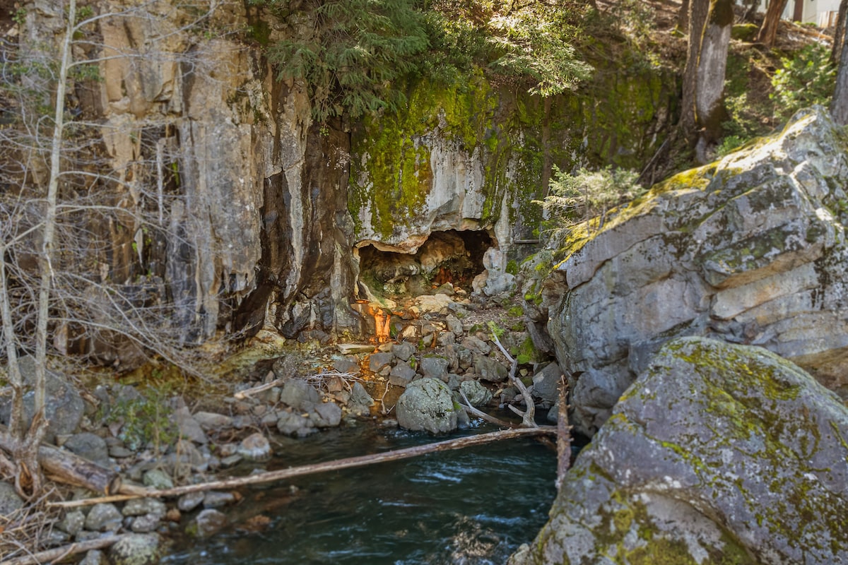 A natural setting features a rocky riverbank beside a gently flowing stream. Moss-covered rocks and vegetation create a serene environment. Sunlight filters through trees, illuminating a small cave situated among the boulders, hinting at the beauty of the nearby landscape.