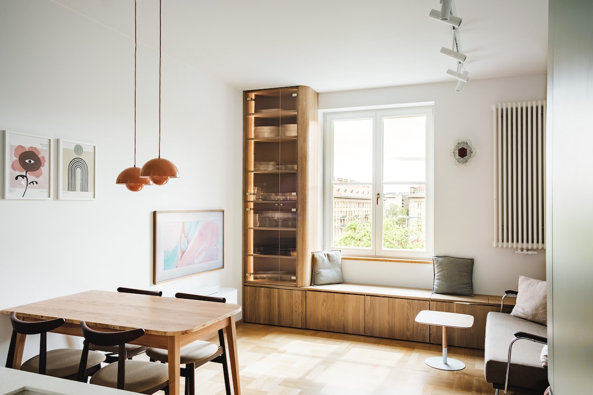 A modern living area is observed, featuring wooden furniture and a comfortable seating arrangement. Large windows allow natural light to flood the space, with views of the surrounding area visible. Decorative art pieces adorn the walls, contributing to the contemporary ambiance.