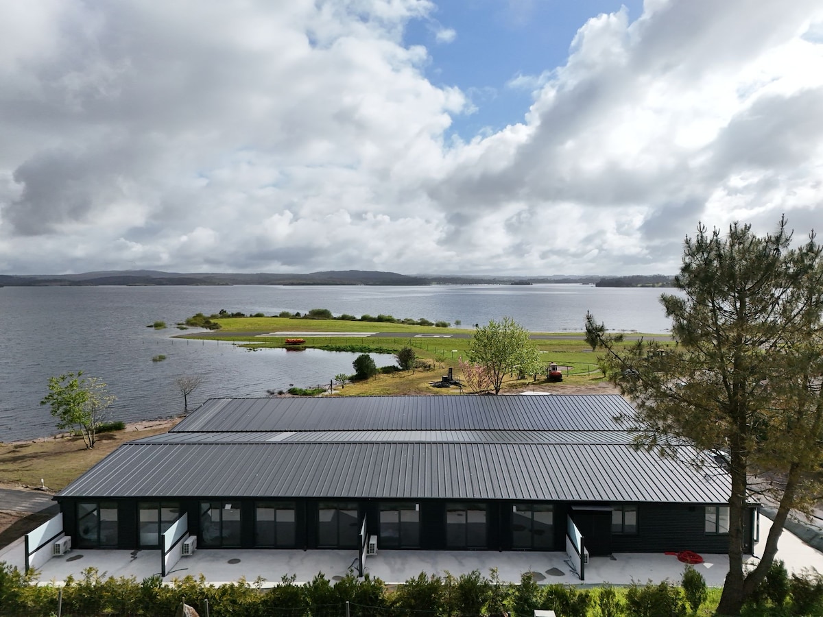 A wide view of the accommodation situated by the lake, featuring a modern building with a dark roof. Lush greenery and a serene landscape surround the water, reflecting the sky's clouds. The tranquil setting is ideal for relaxation in nature.