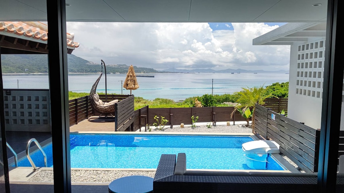 A serene view of a private outdoor pool framed by wooden decking is presented. The pool reflects the sky and surrounding landscape, while a hammock and palm tree evoke a relaxing atmosphere. The backdrop features calm waters and distant mountains under a partly cloudy sky.