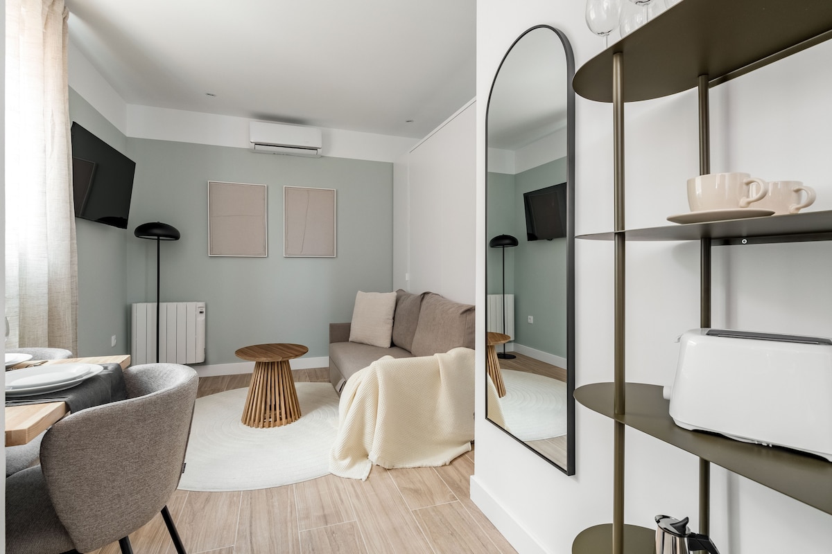 A contemporary living space features a light-toned sofa with accent pillows, paired with a circular wooden coffee table. Two televisions are mounted on the walls, and a round area rug adds texture underfoot. A lean shelf displays kitchenware, while a mirror reflects the room's light.