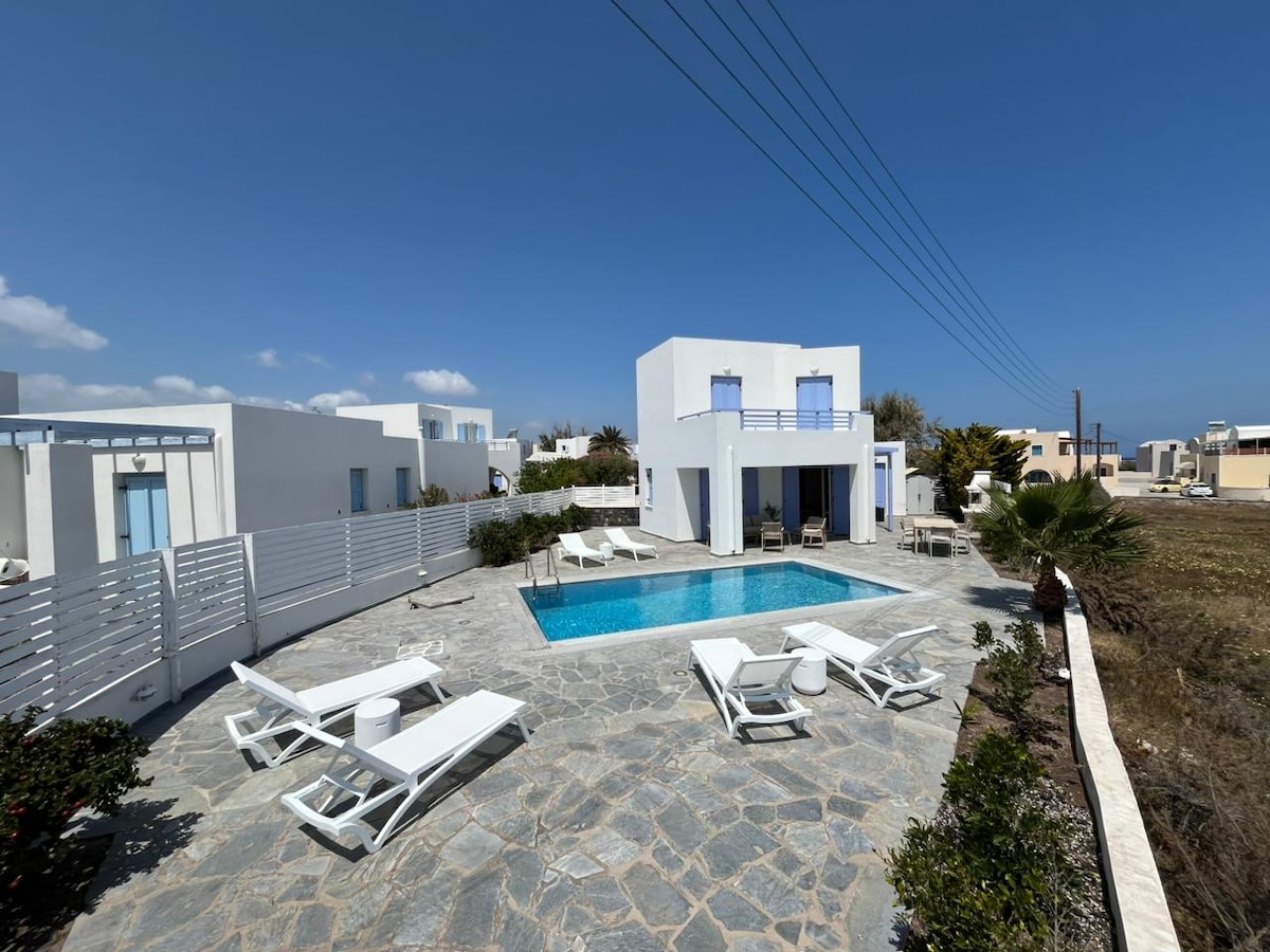 A spacious outdoor area features a large private pool surrounded by sun loungers and small side tables. The villa's bright white exterior and blue window shutters are contrasted against the clear blue sky. Lush greenery borders the stone pathway leading to the pool.