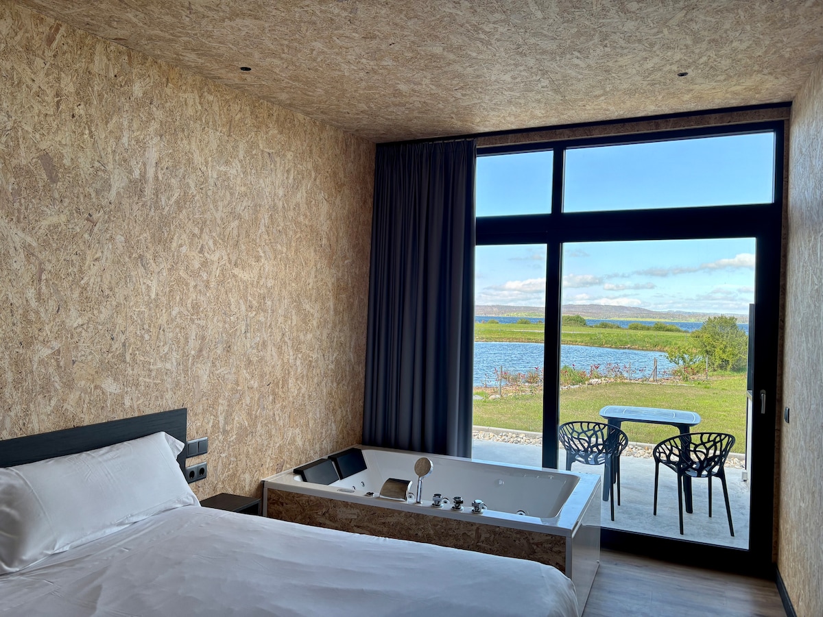 A cozy bedroom features a spacious bed and a Jacuzzi tub, with large windows offering views of the surrounding nature. A small outdoor seating area with two chairs and a table is visible just beyond the glass, allowing for relaxation while enjoying the landscape.