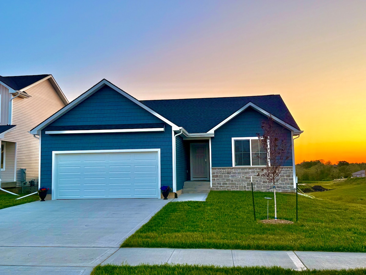 A two-story house is shown with a modern blue exterior featuring light stone accents. A spacious driveway leads to a two-car garage, with manicured grass in the foreground. A sunset creates a warm glow behind the home, enhancing the peaceful setting.