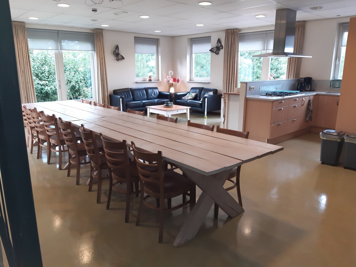 A spacious common area features a long, light-colored wooden table surrounded by multiple wooden chairs, providing ample seating for large gatherings. Comfortable black leather sofas sit in a cozy corner, complemented by a side table. Large windows allow natural light to fill the room.