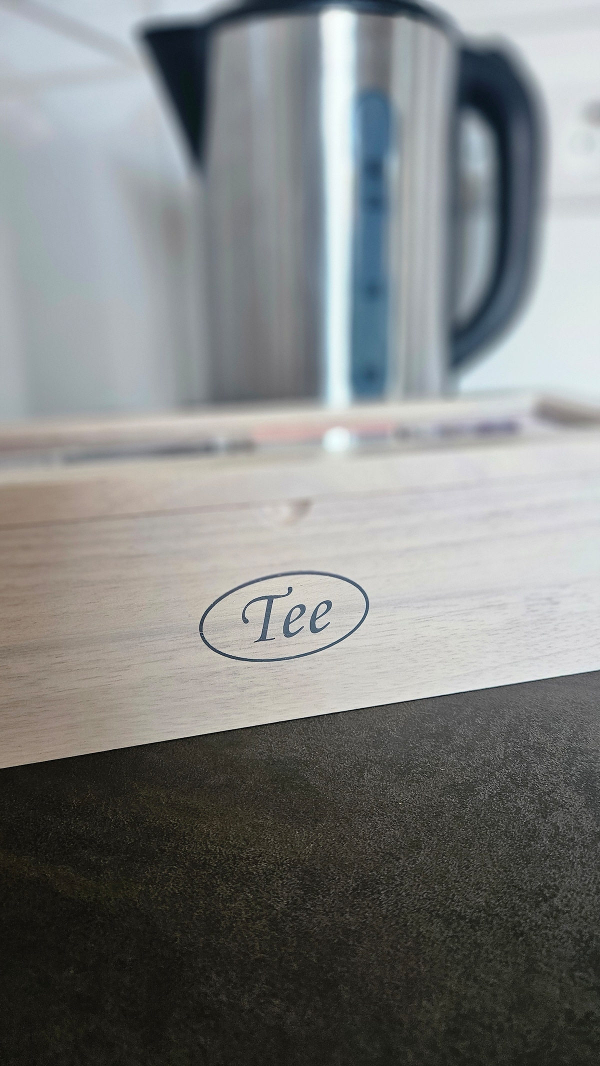A wooden box labeled 'Tee' is placed on a dark countertop, with a sleek metallic kettle slightly blurred in the background. The kettle features a modern design, complementing the warm tones of the wood.