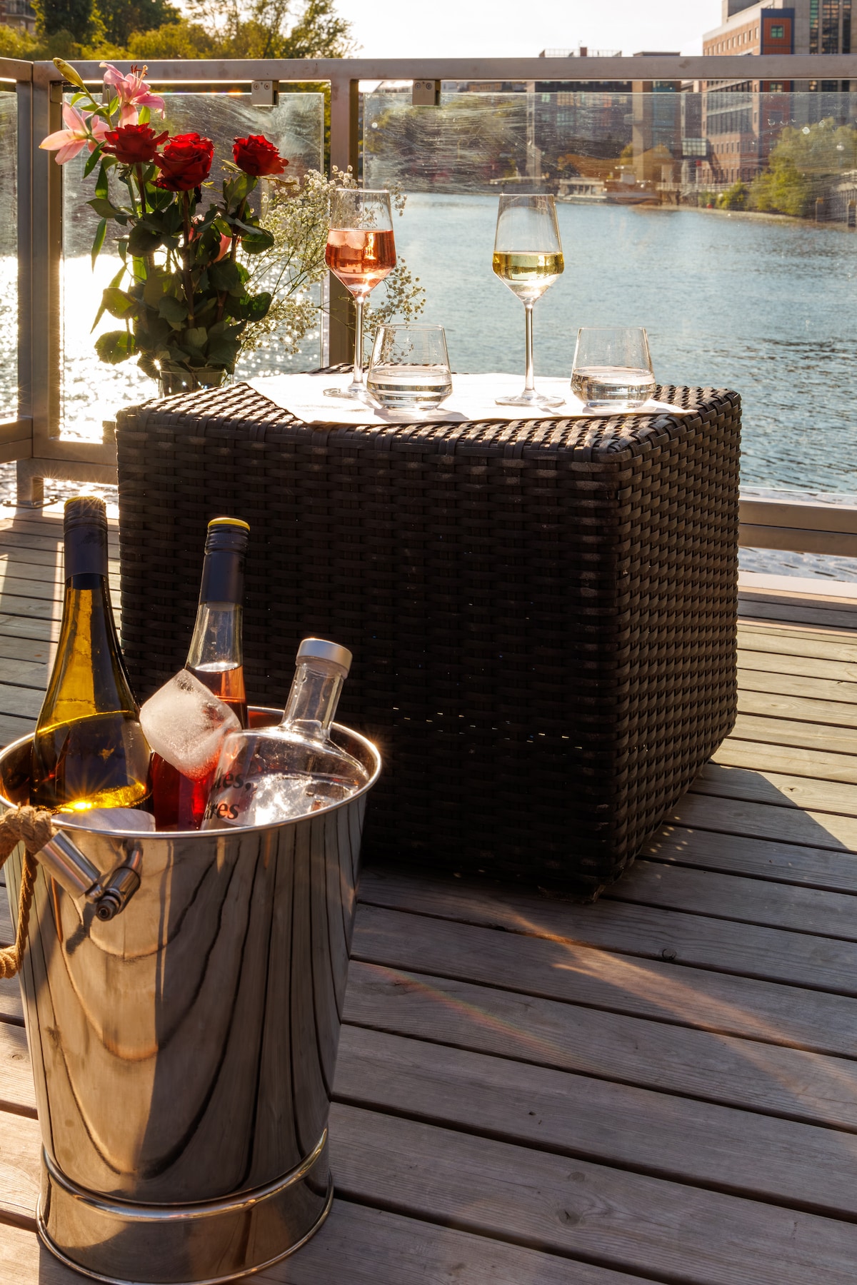 A small table is set on a wooden deck, accompanied by four glasses and two bottles of wine in an ice bucket. A bouquet of flowers adds a touch of color against the backdrop of the water and nearby buildings.