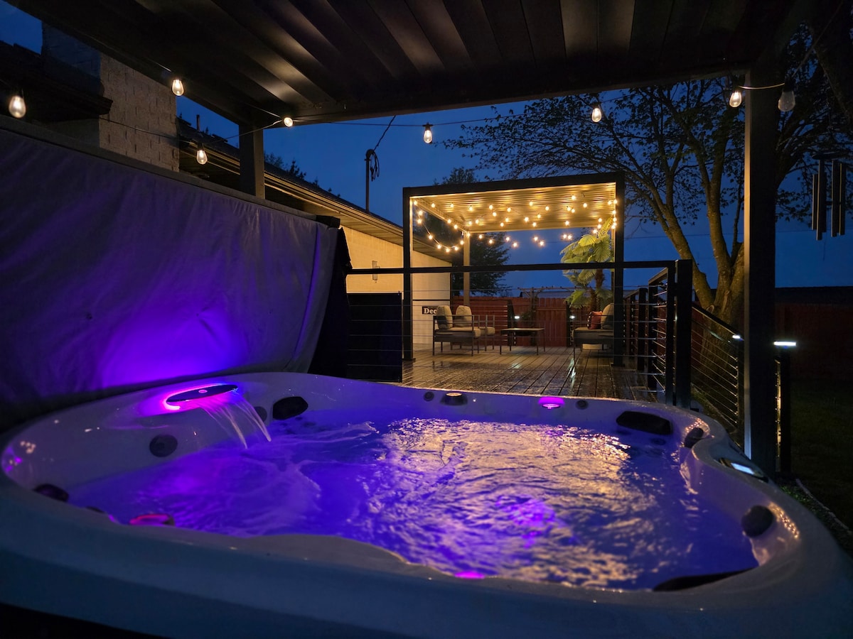 A private hot tub bubbles gently under a softly lit pergola, illuminated by string lights. The evening sky provides a calm backdrop, while a spacious deck with seating options invites relaxation and enjoyment.