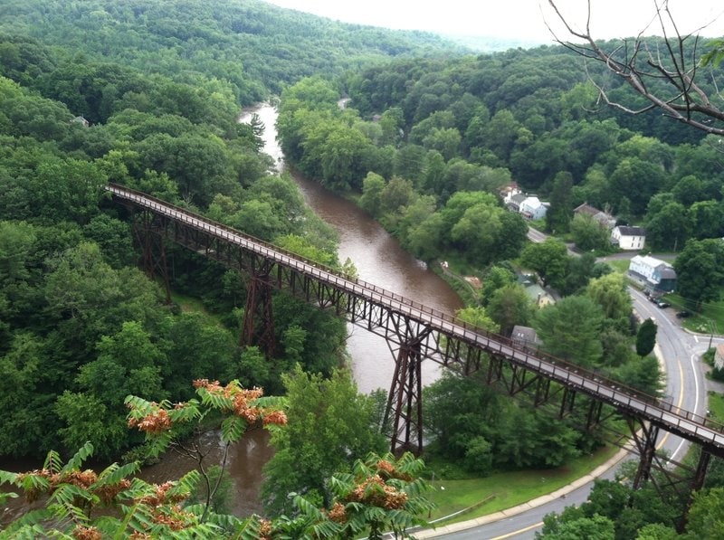 A high-angle view displays a lengthy trestle bridge spanning a winding river, surrounded by lush green foliage. The landscape features gentle slopes and a mix of trees, with a few houses visible along the riverbank. A road curves near the river, adding to the scenic view.