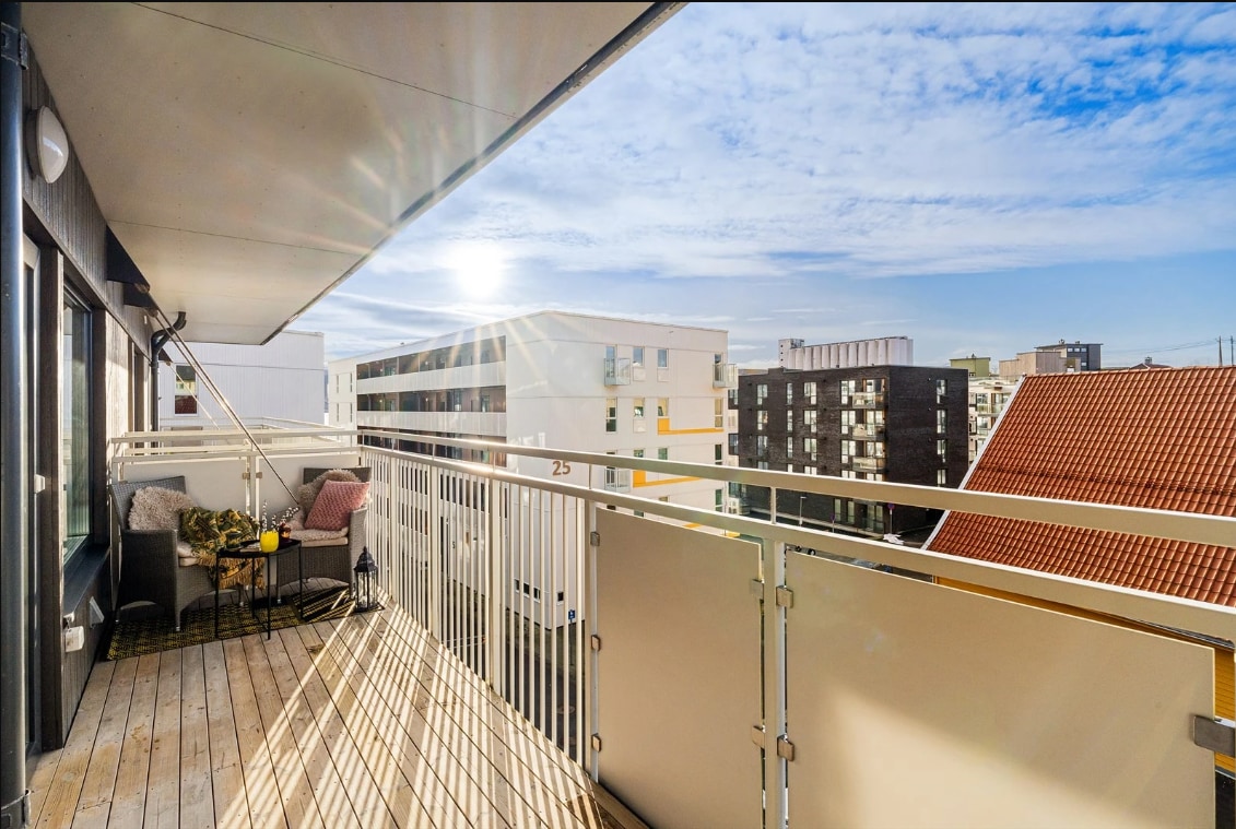 A balcony features wooden flooring and a metal railing, providing views of surrounding buildings and the sky. Sunlight bathes the space, creating a bright atmosphere. A small seating area is visible, including two chairs and a table, inviting relaxation.