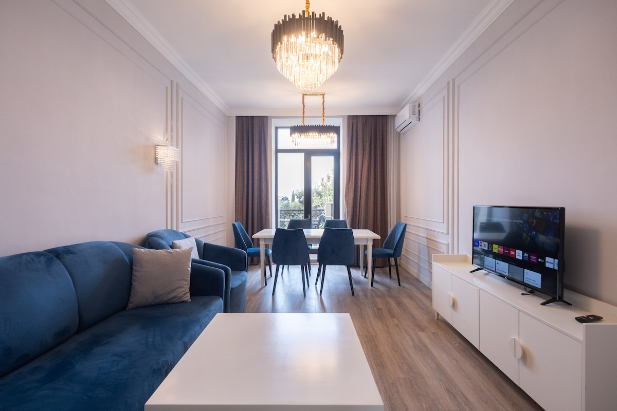 A spacious living area features a blue sofa paired with matching armchair, complemented by a modern chandelier overhead. A white coffee table is positioned centrally, and a large flat-screen TV is set against a light-colored media unit. Natural light filters through large windows leading to one of the balconies.
