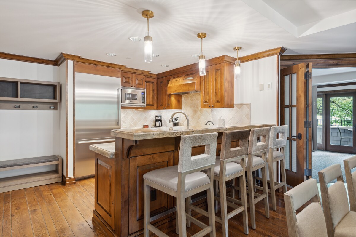 An updated gourmet kitchen features warm wooden cabinetry and stone countertops. A spacious bar with four stools invites casual dining. Stainless steel appliances are visible, complemented by soft, recessed lighting that enhances the overall inviting environment.