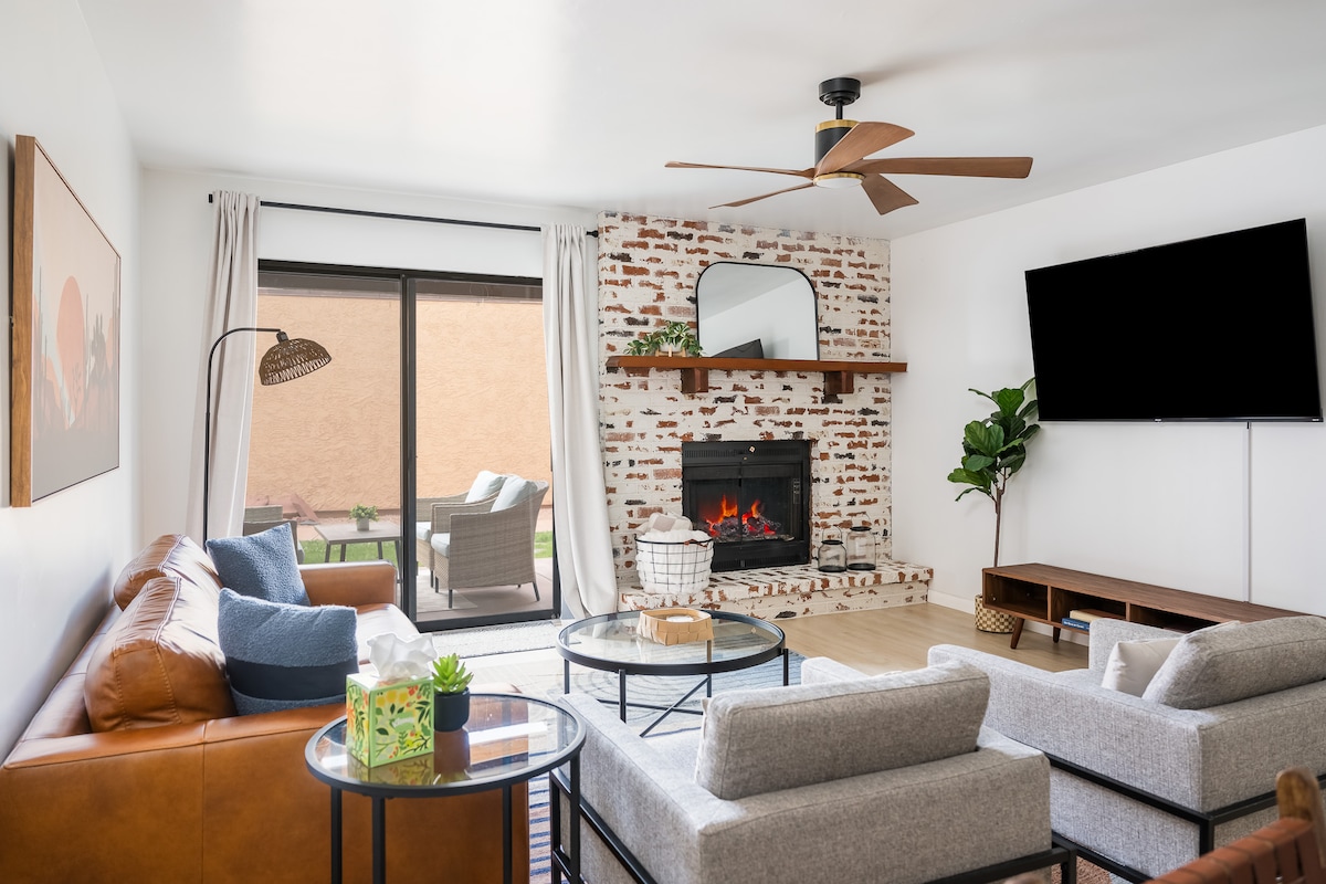 A cozy living room features a blend of comfortable seating, including a leather sofa and light gray chairs. A brick fireplace with a mantel is highlighted, while natural light pours in through sliding glass doors that lead to a patio. A large television is mounted on the wall.