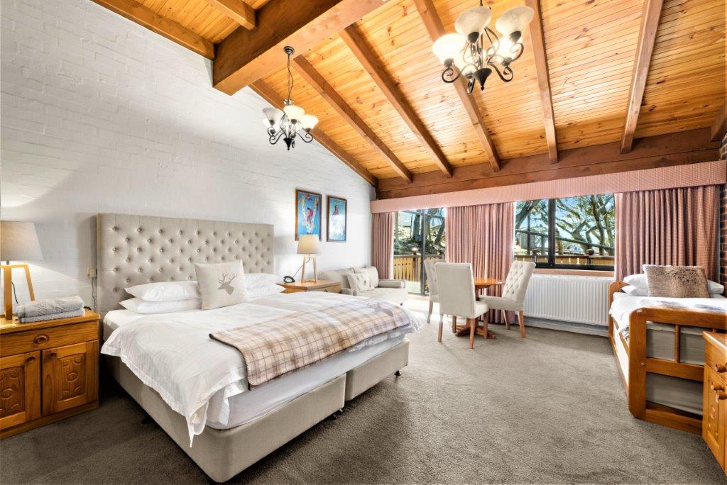 A spacious hotel room features a king bed and two single beds with soft bedding. Rich timber beams enhance the ceiling, while large windows with sheer curtains provide natural light. A cozy seating area with a table and two chairs overlooks the balcony.
