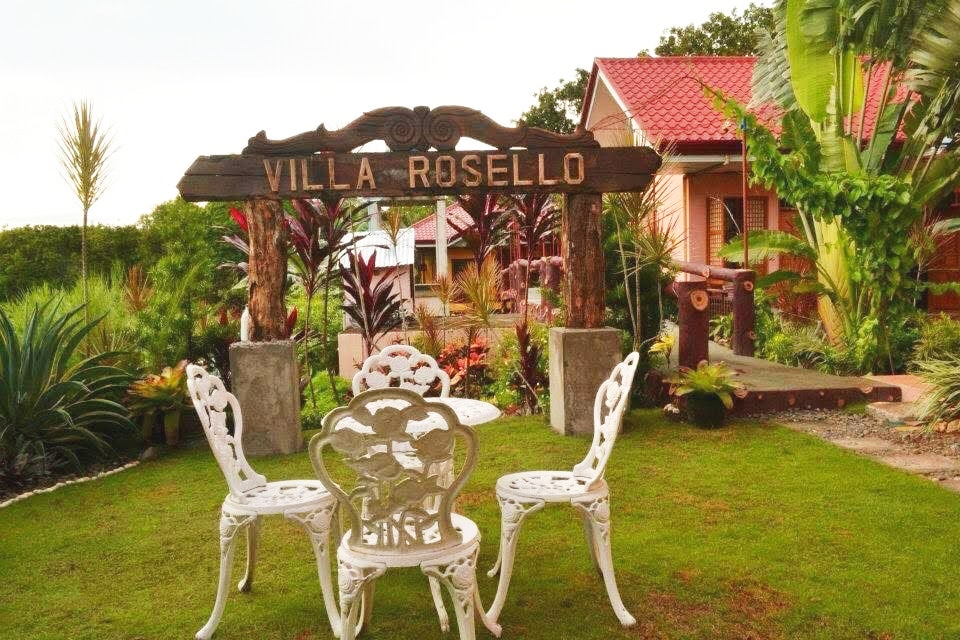 A wooden sign reading 'Villa Rosello' welcomes guests at the entrance, flanked by vibrant tropical plants. A quaint seating area with white metal chairs and a round table sits on lush grass, inviting relaxation amidst the picturesque gardens.