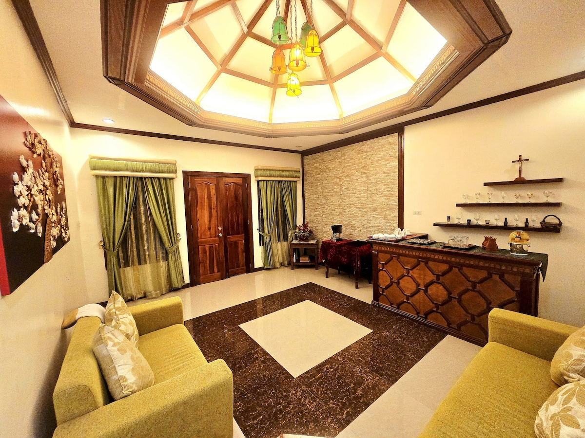 A spacious lounge area features two green sofas positioned across from a wooden reception counter. Soft lighting is provided by a decorative ceiling fixture, while the walls are adorned with greenery-inspired drapes. A stone accent wall adds texture to the room.