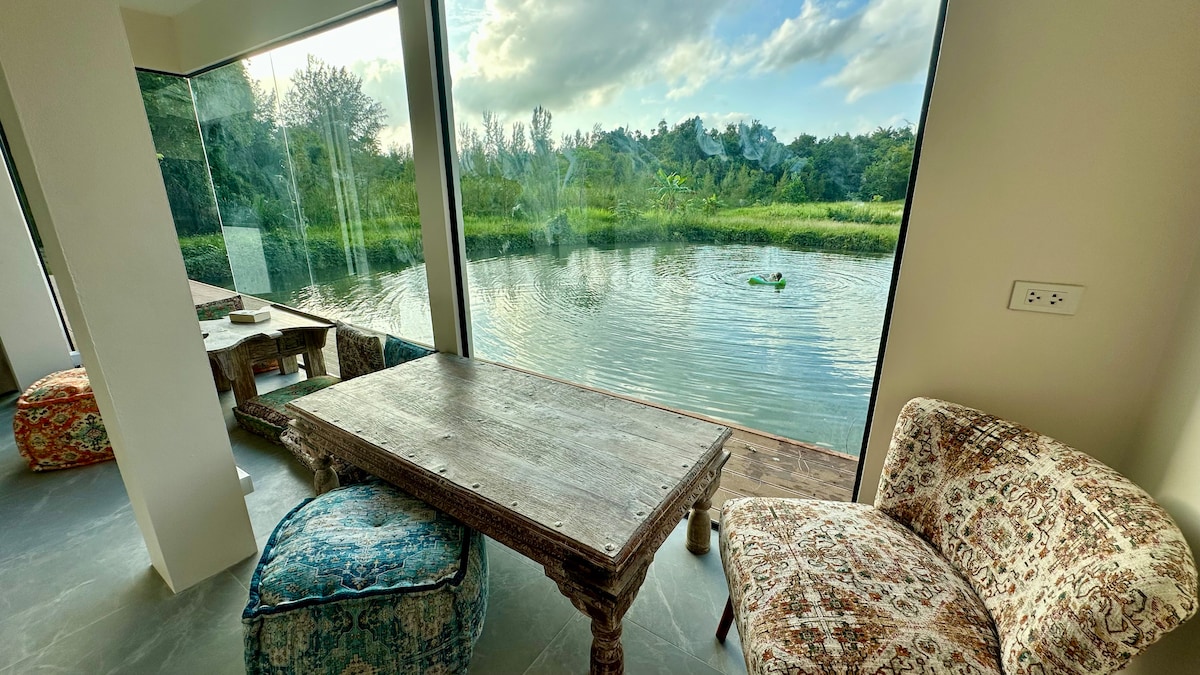 A tranquil seating area features a rustic wooden table surrounded by patterned chairs and poufs. Large glass windows provide a clear view of the serene lake and lush greenery just beyond, inviting natural light into the space.