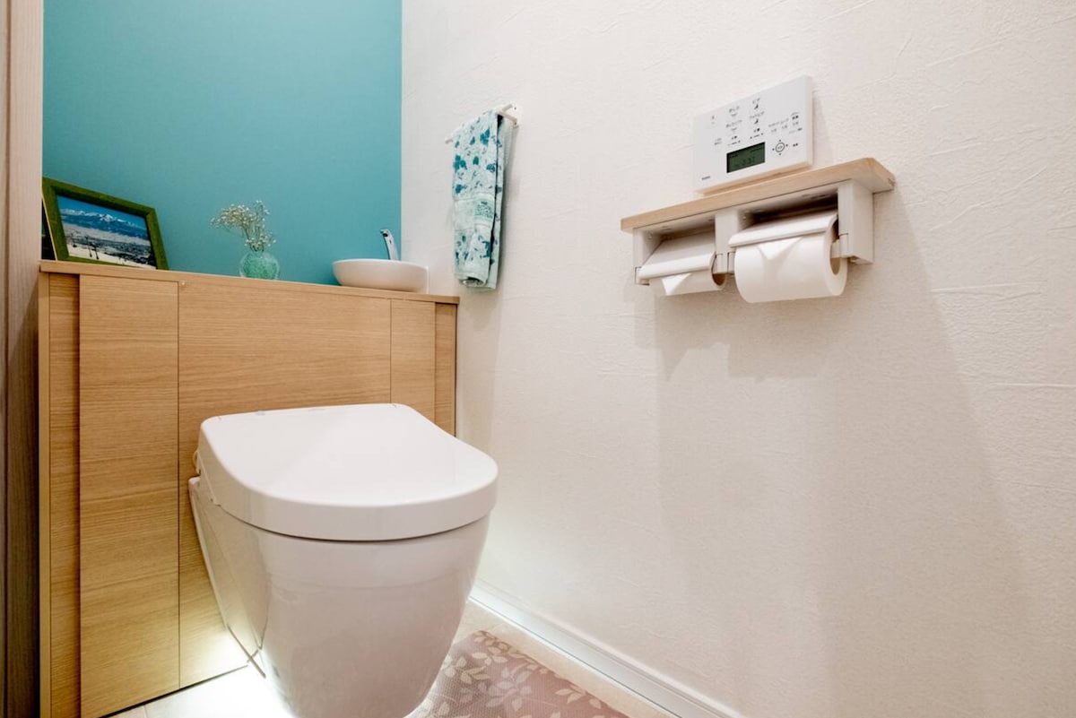 A modern bathroom is depicted, featuring a sleek toilet accompanied by wooden cabinetry. A small basin is positioned on the counter, with a decorative towel hanging nearby. Soft lighting creates a calm environment, complemented by a light-colored rug on the floor.