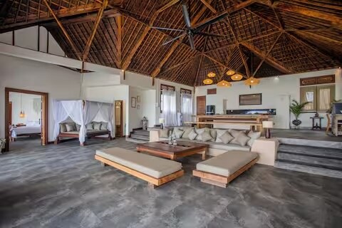 The open-plan living area features a high, intricately designed wooden ceiling with hanging lights. A large, low wooden table is surrounded by spacious, cushioned seating, creating a relaxed atmosphere. Views into the bedrooms and kitchen area are visible through wide openings in the layout.