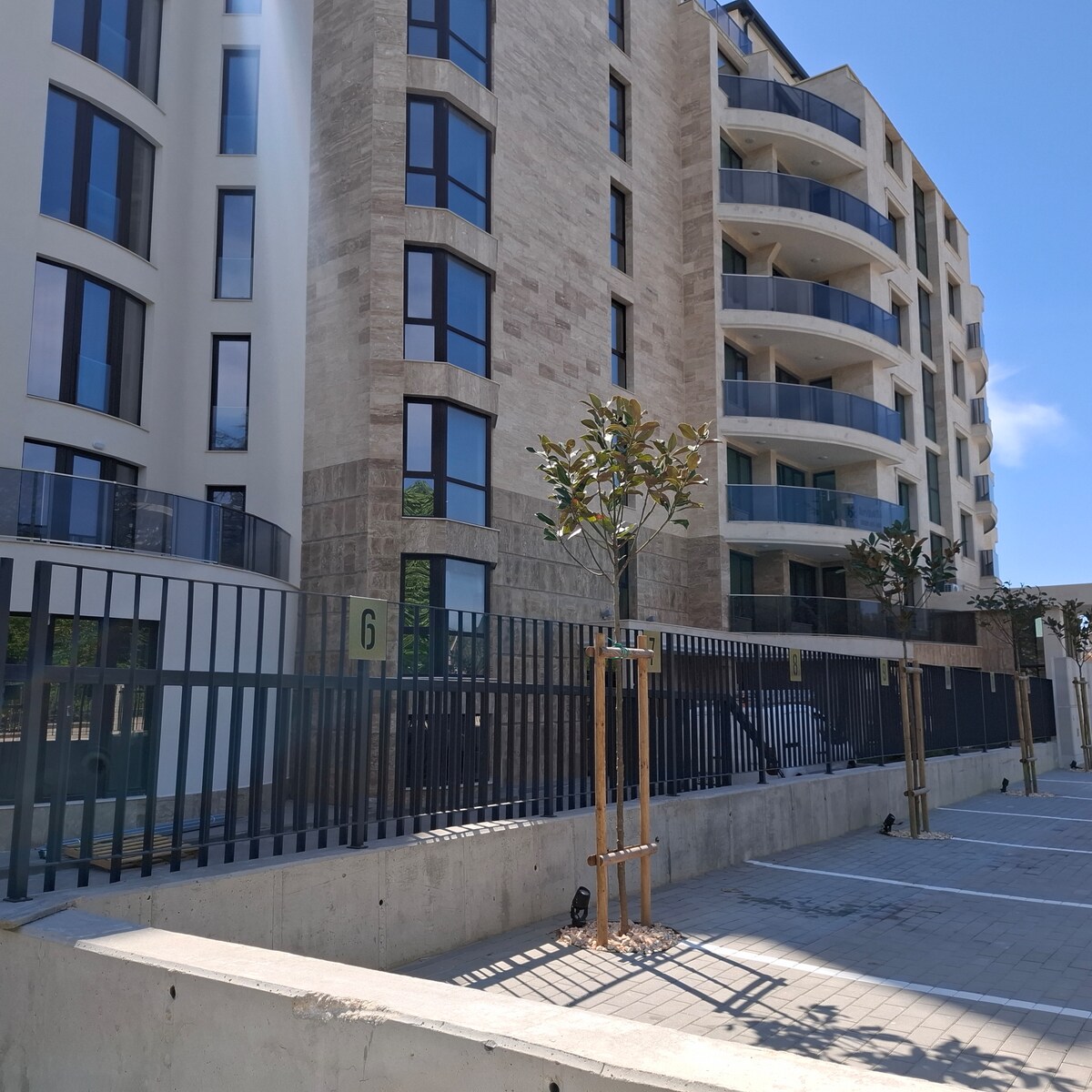 The exterior of a contemporary apartment building is showcased, featuring a combination of stone and glass elements. Large windows and rounded balconies provide a modern aesthetic. Newly planted trees line the entrance, adding greenery to the urban landscape, while the spacious paved area is visible in the foreground.
