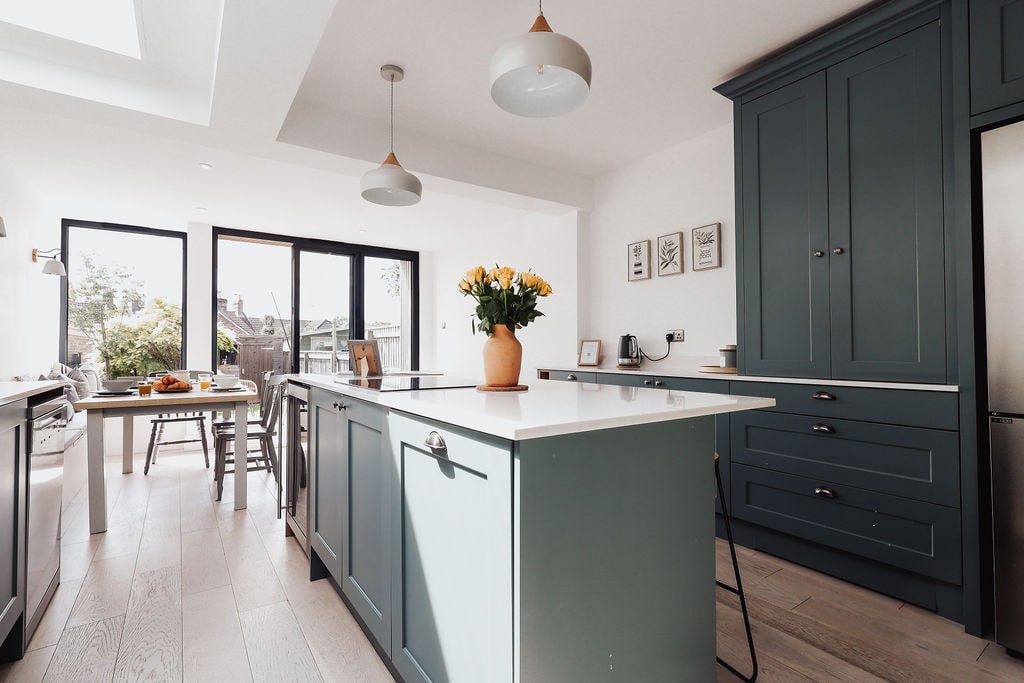 A modern kitchen is depicted, featuring a central island with a white countertop and blue cabinetry. Large windows allow ample natural light to enter the space, and a vase of yellow flowers adds a touch of color. A dining table is set in the background, indicating a welcoming atmosphere.