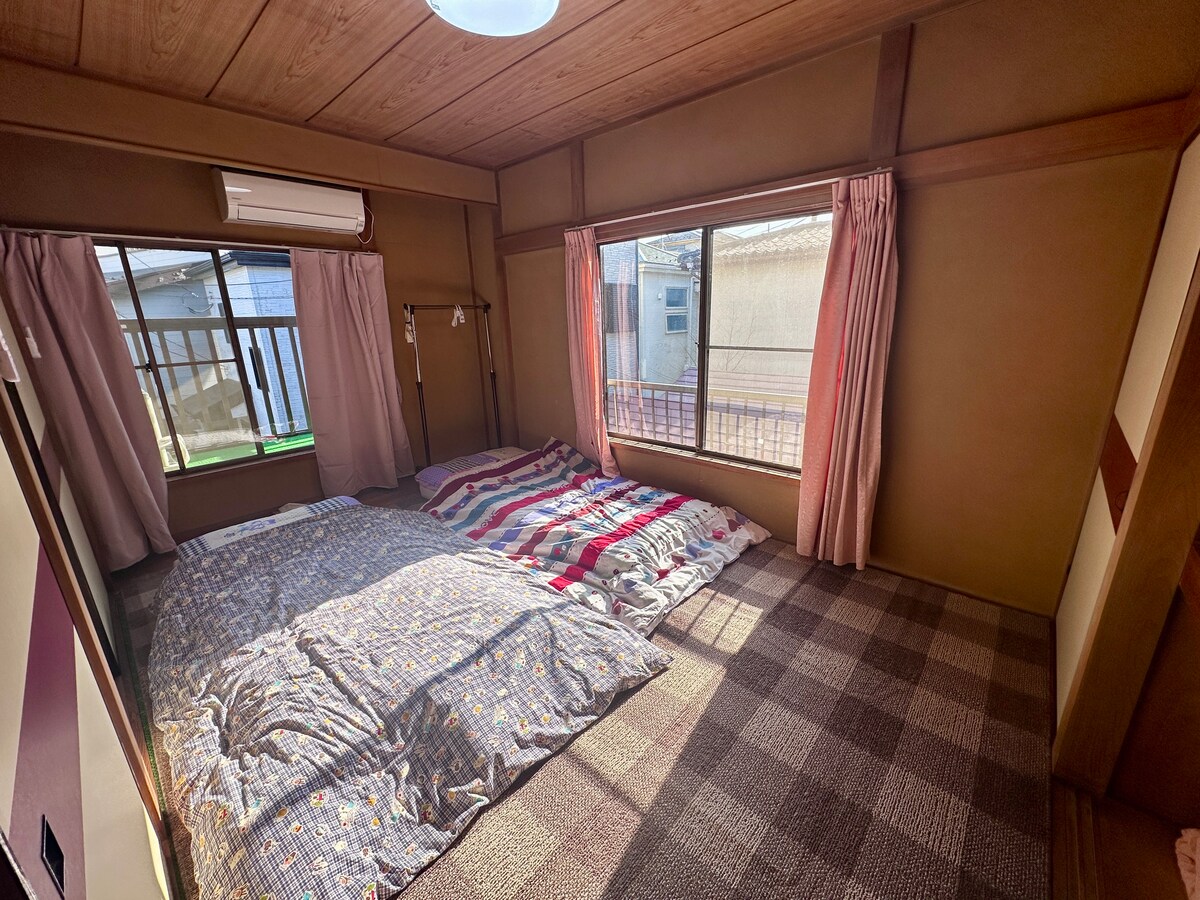 A traditional Japanese room features two futons arranged on a textured tatami mat. Light-colored curtains frame the large windows, allowing natural light to fill the space. A serene ambiance is created by the warm wooden walls and ceiling, complemented by a minimalist design.